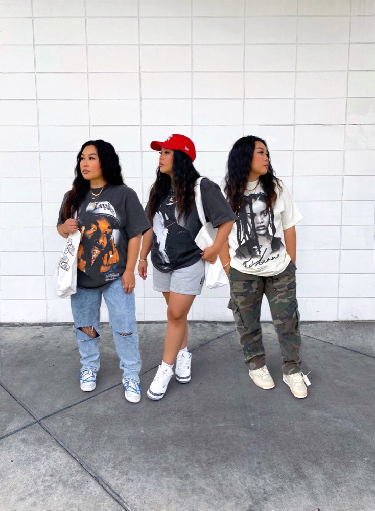 Three women in graphic tees standing side by side against a white tile wall, looking to the side
