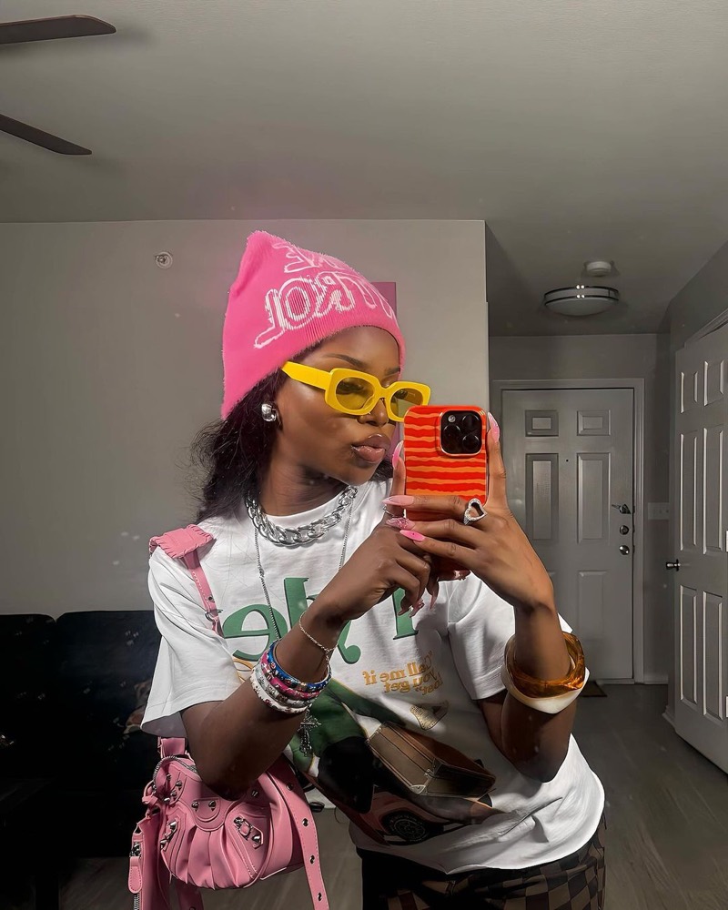 An influencer taking a mirror selfie in a pink beanie, yellow sunglasses and a graphic tee