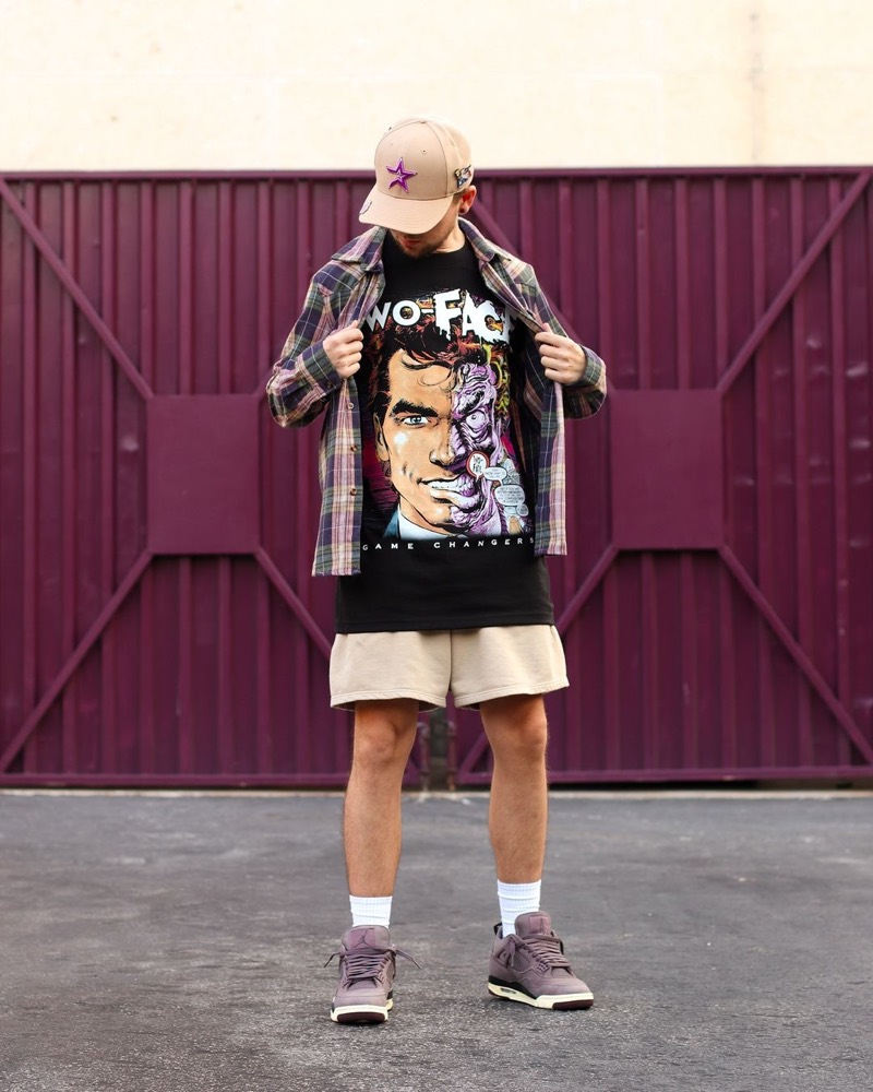A man in a cap and a plaid shirt opened over a graphic tee, in front of a maroon gate