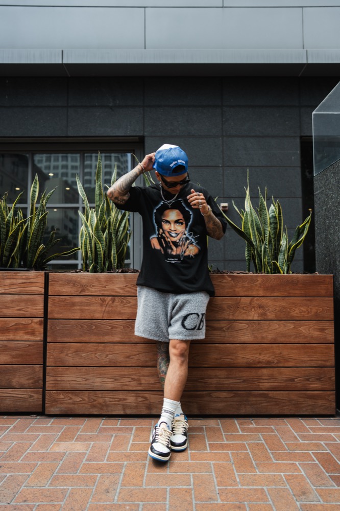 A man in a blue trucker cap and sunglasses, looking down, in a black graphic tee and grey knit shorts on a city street