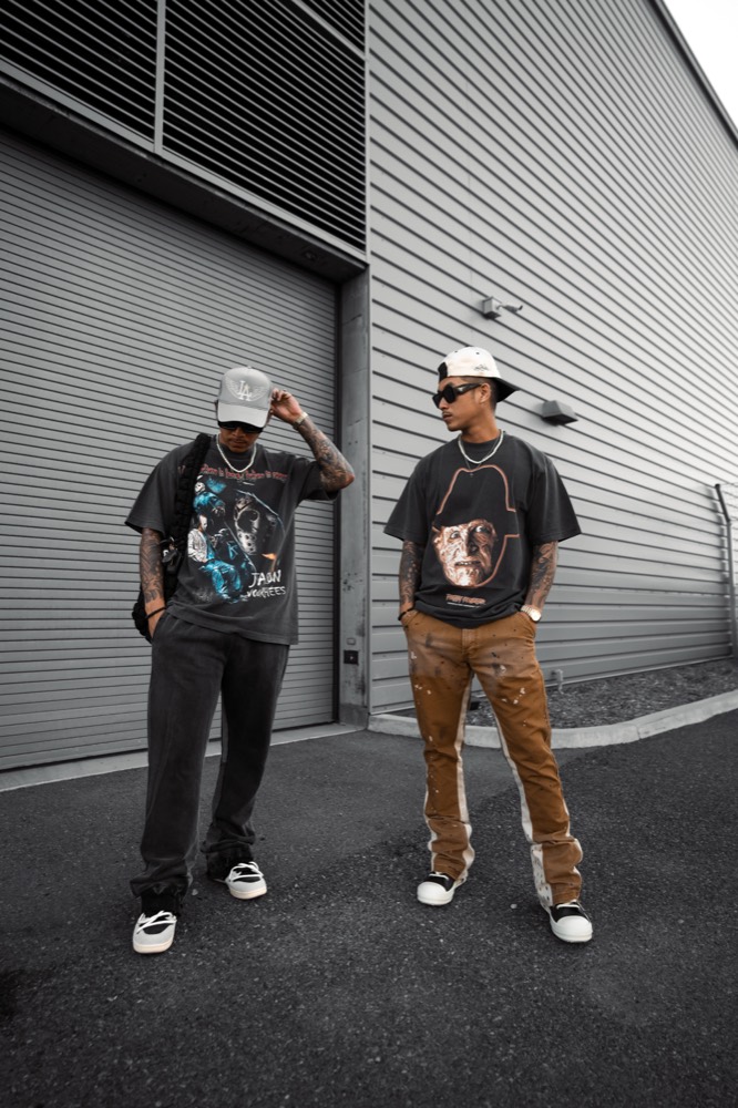 Two men in graphic tees standing side by side in front of a grey warehouse