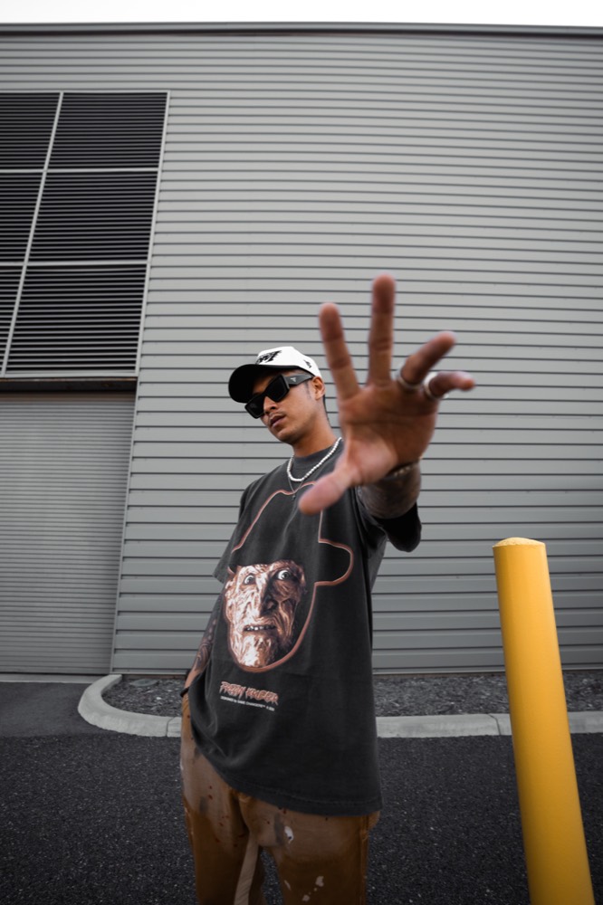 A man reaching a hand toward the camera in a white cap, sunglasses and a graphic tee, against a grey warehouse wall