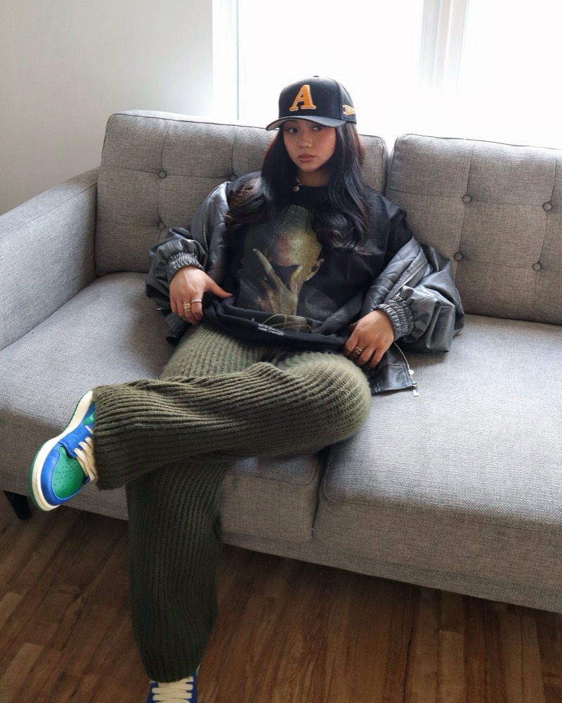 A woman on a grey sofa in a black cap, a graphic tee and olive knit pants