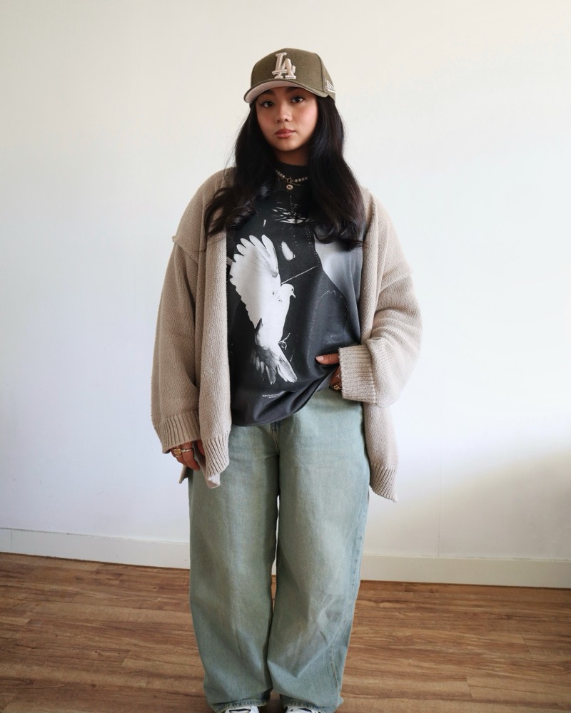 A woman in an olive cap, a beige cardigan, a graphic tee and wide jeans against a white wall