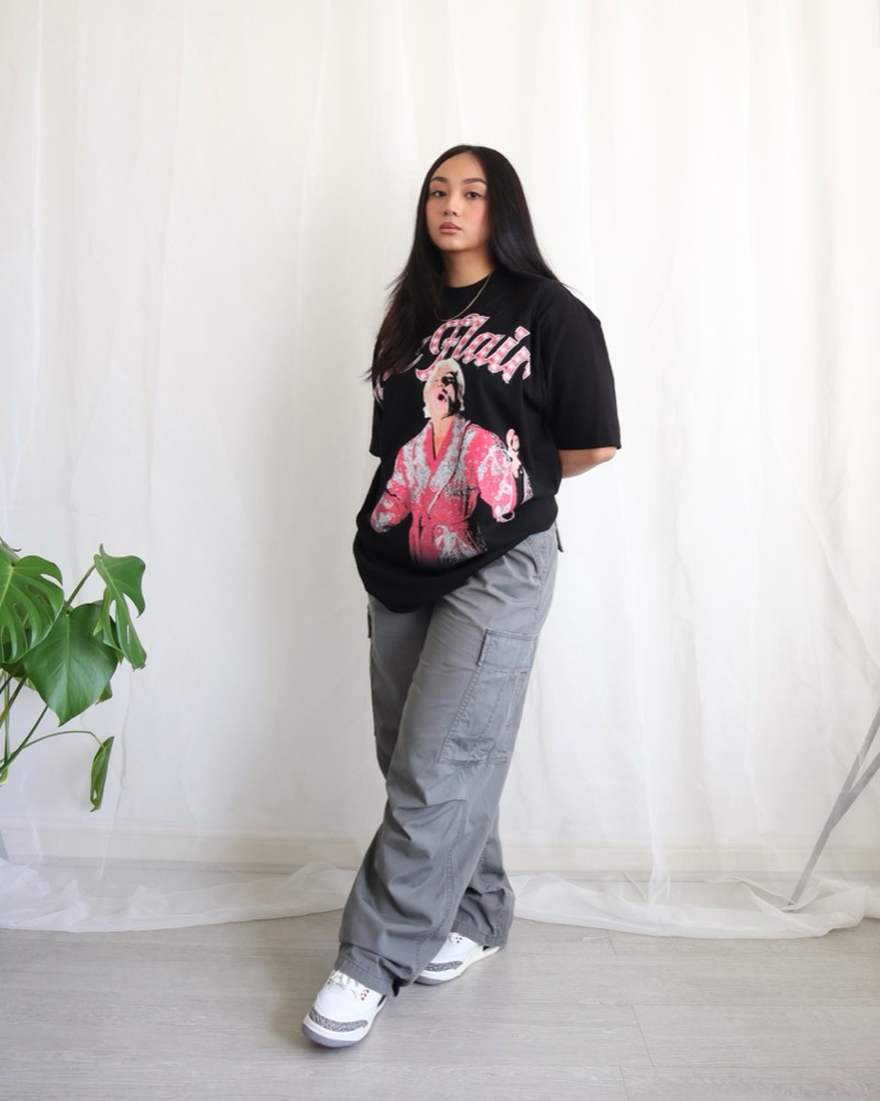 A woman in a black graphic tee and grey cargo pants, leaning in front of sheer white curtains