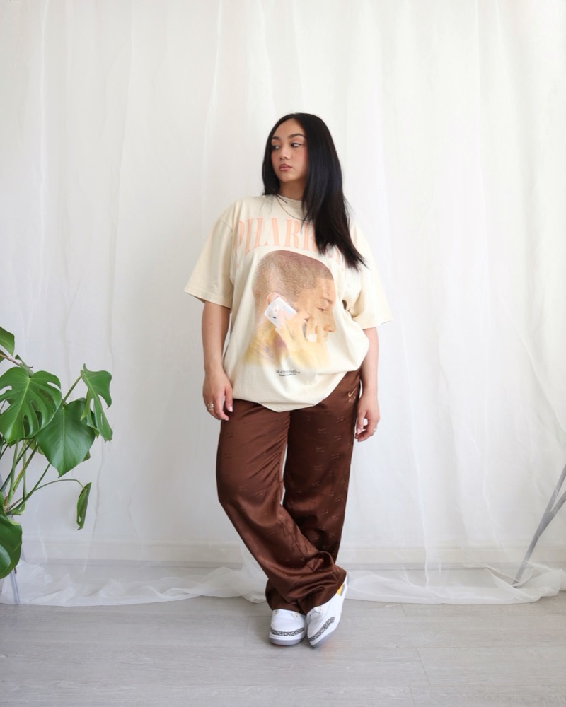 A woman in a cream oversized graphic tee and brown satin pants against sheer white curtains