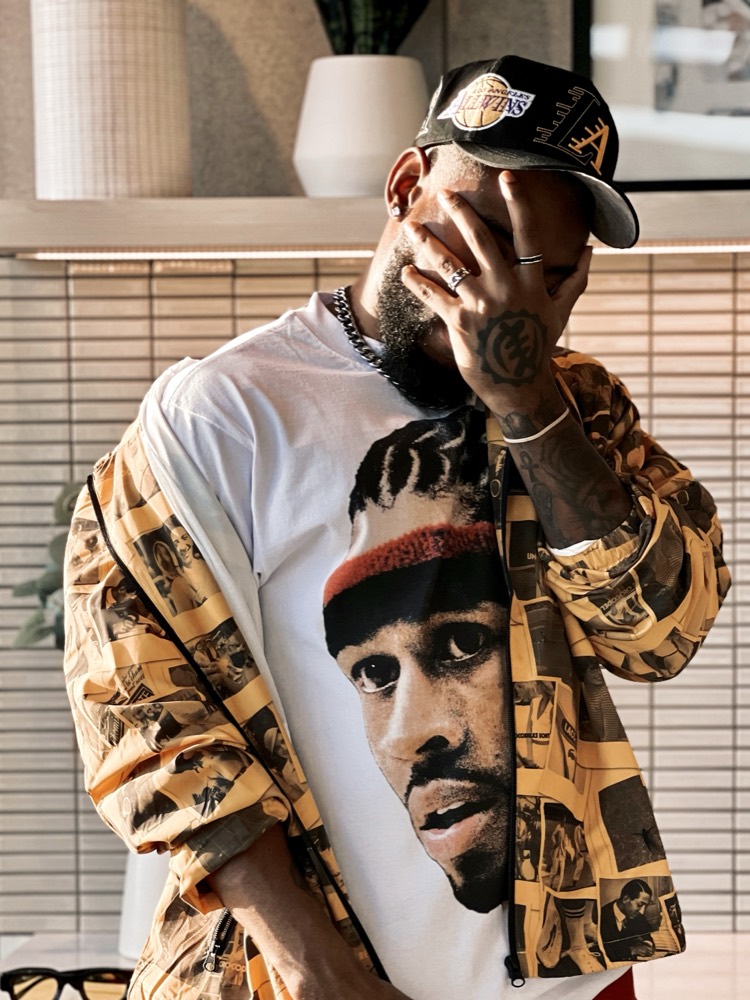 A man in a Lakers cap and a patterned jacket over a graphic tee, with a hand over his face