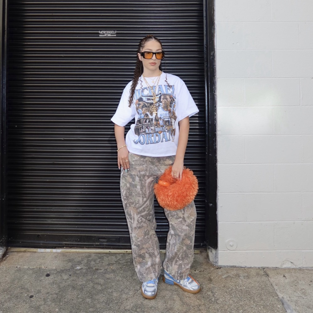 A woman in orange sunglasses and a graphic tee, holding an orange fur bag, in front of a dark garage door